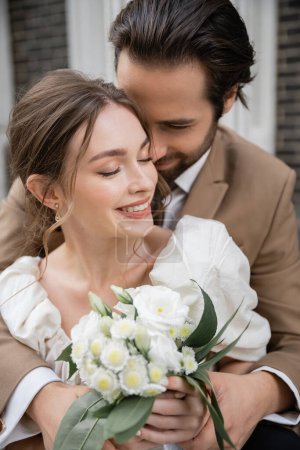 bearded groom hugging happy bride in white dress with wedding bouquet 