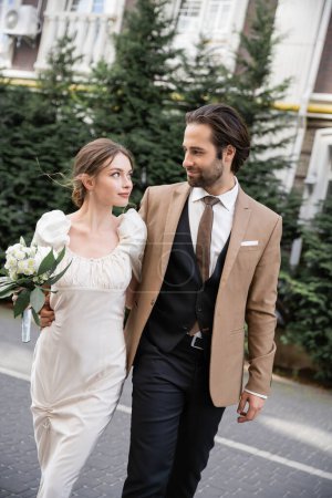 young bride in white dress holding wedding bouquet while walking with bearded groom  on street 