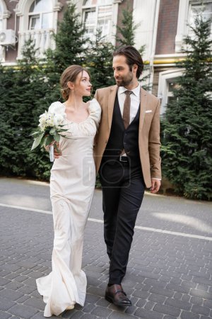 full length of gorgeous bride in white dress holding wedding bouquet while standing with bearded groom on street 