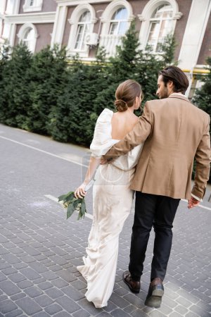 back view of bride in white dress holding wedding bouquet and walking with groom on street 