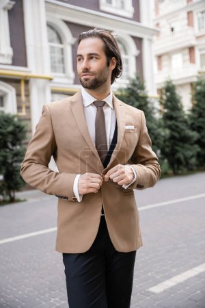 young groom in beige formal wear buttoning blazer on street 