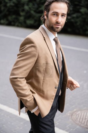 young groom in beige formal wear standing with hand in pocket and looking at camera 
