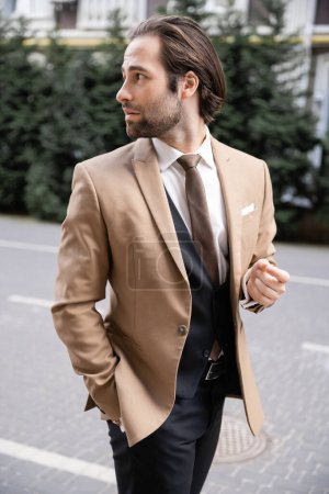 novio guapo en ropa formal beige de pie con la mano en el bolsillo y mirando hacia otro lado