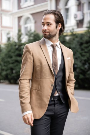 young groom in beige formal wear standing with hand in pocket and looking away on street 
