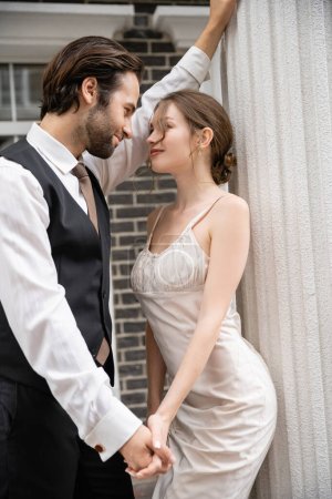 Foto de Groom in formal wear and bride in wedding dress holding hands while looking at each other - Imagen libre de derechos