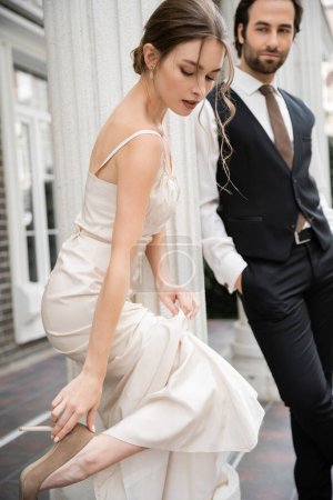 pretty bride in wedding dress wearing high heeled shoe near groom on blurred background 