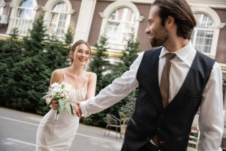 happy bride in white dress with wedding bouquet holding hands with groom while walking outside 
