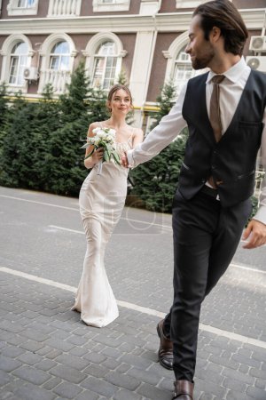 full length of groom in vest holding hand of bride in white dress with wedding bouquet 