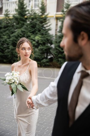 blurred groom in vest holding hand of gorgeous bride in white dress with wedding bouquet 