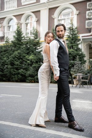 full length of young bride and groom in wedding dress and suit holding hands outside 