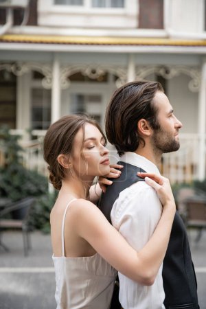 young bride in wedding dress standing with closed eyes and hugging groom in vest 