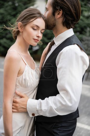 bärtiger Bräutigam in Weste und Hemd umarmt Braut im weißen Hochzeitskleid 