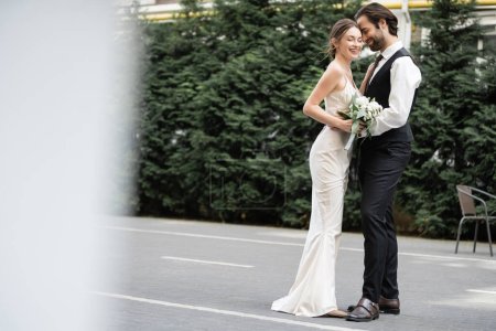 Téléchargez les photos : Happy groom in vest embracing cheerful bride in white dress with wedding bouquet - en image libre de droit