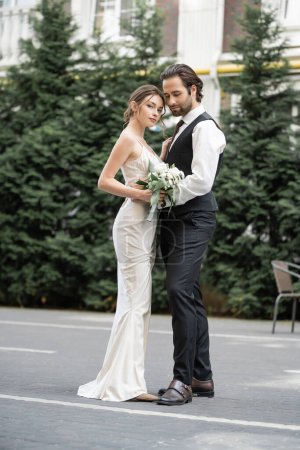 Téléchargez les photos : Full length of bearded man in vest embracing bride in white dress with wedding bouquet - en image libre de droit