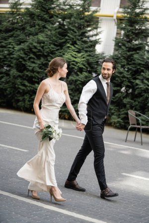 longitud completa del novio sosteniendo la mano de la novia en vestido blanco con ramo de flores 