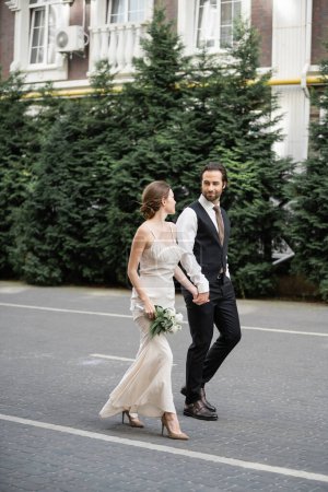 longitud completa del hombre en traje de la mano de la novia en vestido blanco con ramo de flores 