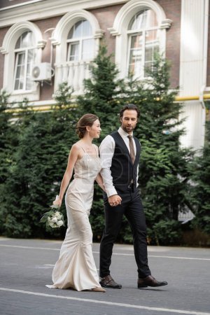 longitud completa del novio caminando con novia feliz en vestido blanco sosteniendo ramo de flores 