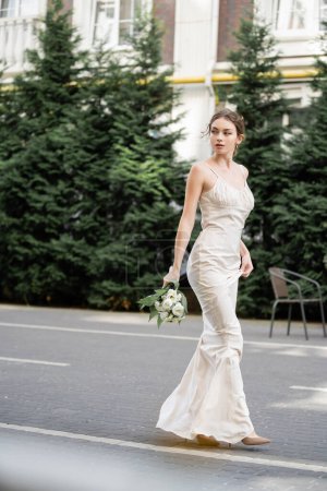 full length of young woman in white dress holding wedding bouquet of blooming flowers and standing outside 