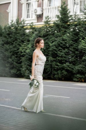 full length of young woman in white dress holding wedding bouquet of flowers and standing outside 