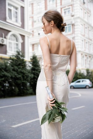 pretty young woman in white dress holding wedding bouquet behind back 