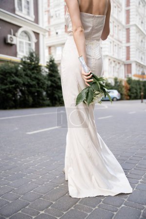 partial view of young woman in white dress holding wedding bouquet behind back 