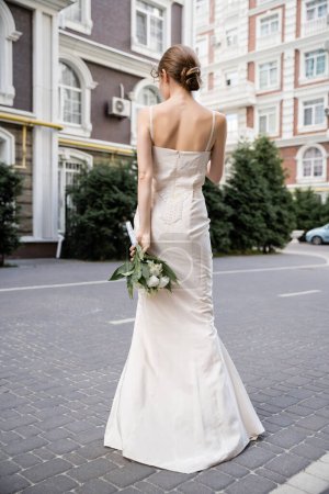 back view of young woman in white dress holding wedding bouquet behind back 