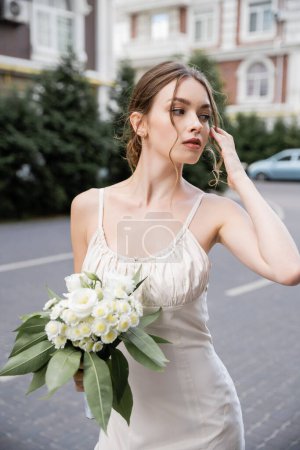 young woman in wedding dress holding bouquet with flowers and looking away