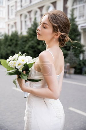 jeune mariée en robe blanche debout avec les yeux fermés et tenant bouquet de mariage 