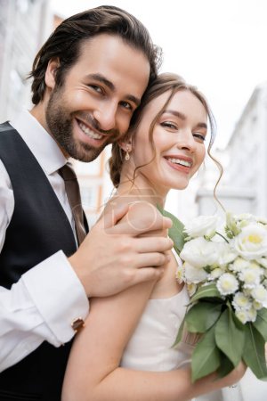 portrait of happy groom with beard hugging cheerful bride holding wedding bouquet 