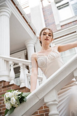 low angle view of young bride in wedding dress looking at camera near house 