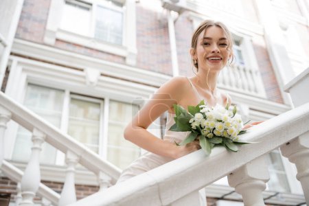 low angle view of happy bride in wedding dress holding bouquet and looking at camera near house 