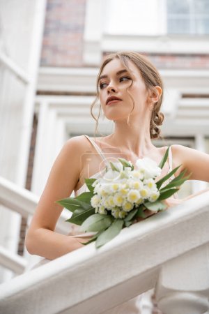 low angle view of bride in wedding dress holding bouquet and looking away near house 