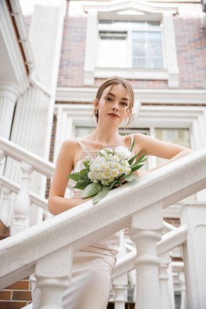 low angle view of bride in white dress holding bouquet and looking at camera near house 