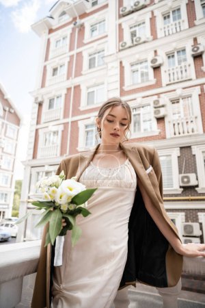 young woman in wedding dress and beige blazer holding bouquet of flowers 