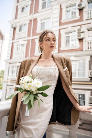 pretty young woman in wedding dress and beige blazer holding bouquet of flowers 