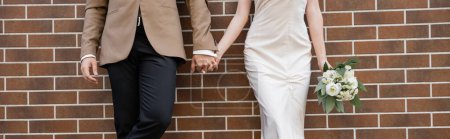 Téléchargez les photos : Cropped view of young bride in white dress holding wedding bouquet and hand of groom near brick wall, banner - en image libre de droit