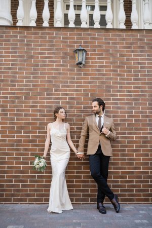 full length of bride in wedding dress holding flowers and hand of groom while standing against brick wall 