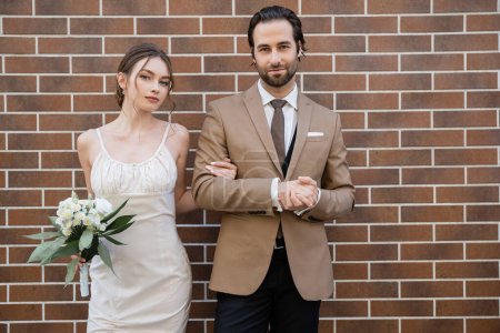Téléchargez les photos : Mariée en robe de mariée tenant des fleurs près du marié en costume tout en se tenant contre le mur de briques - en image libre de droit