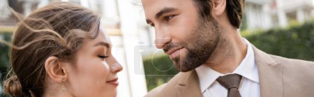 bearded man in suit looking at smiling bride outdoors, banner 