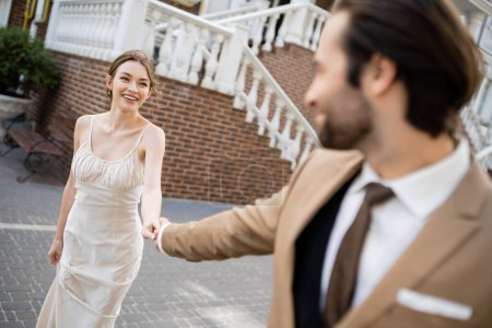 blurred groom in suit holding hand of happy bride in white dress 