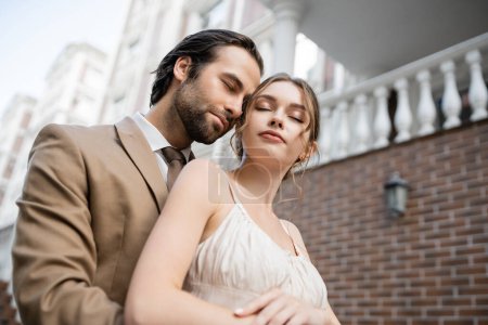 low angle view of groom in beige suit standing near gorgeous bride with closed eyes