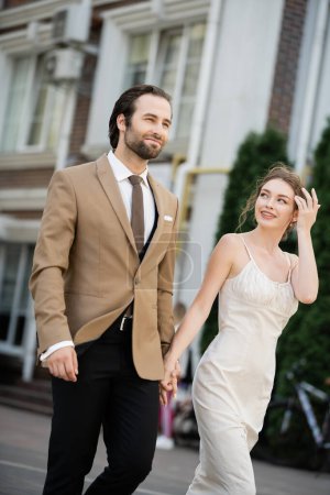 happy bride in wedding dress looking at groom while holding hands on street 