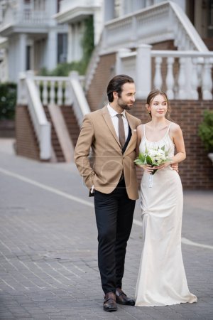 longitud completa del novio posando con la mano en el bolsillo mientras mira a la novia en vestido de novia con flores en flor 