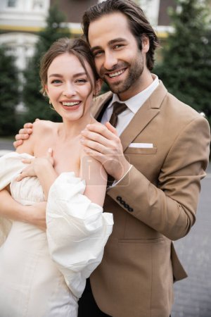 retrato de hombre alegre y barbudo abrazando novia feliz en vestido de novia 