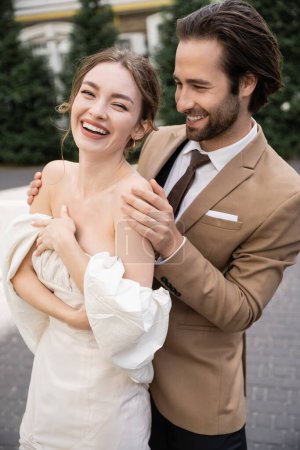retrato de hombre alegre y barbudo abrazando novia positiva en vestido de novia 