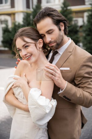 portrait of bearded young man hugging happy bride in white wedding dress 