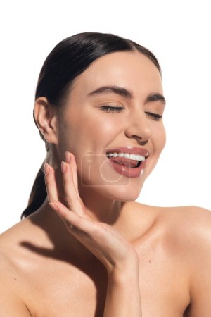 cheerful young woman with natural makeup and bare shoulder touching cheek while sticking out tongue isolated on white