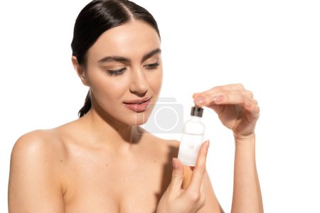 brünette Frau mit nackten Schultern schaut auf Flasche mit feuchtigkeitsspendendem Serum isoliert auf weiß 