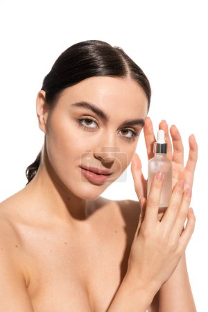 brunette woman with bare shoulders looking at camera while holding bottle with moisturizing serum isolated on white  mug #642937156
