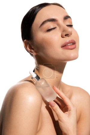 brunette woman with bare shoulders and closed eyes holding bottle with moisturizing serum isolated on white 